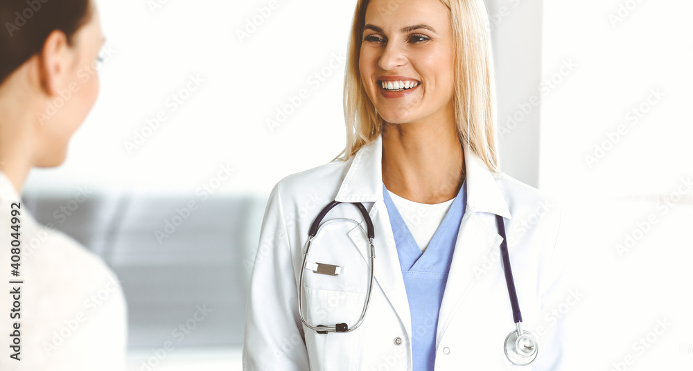 Woman-doctor at work in sunny hospital is happy to consult female patient. Blonde physician checks medical history record and exam results while using clipboard Woman-doctor at work in sunny hospital is happy to consult female patient. Blonde physician checks medical history record and exam results while using clipboard