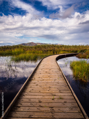 Camino de madera que se adentra en una laguna con vegetación verde y montañas al fondo con un cielo de nubes blancas