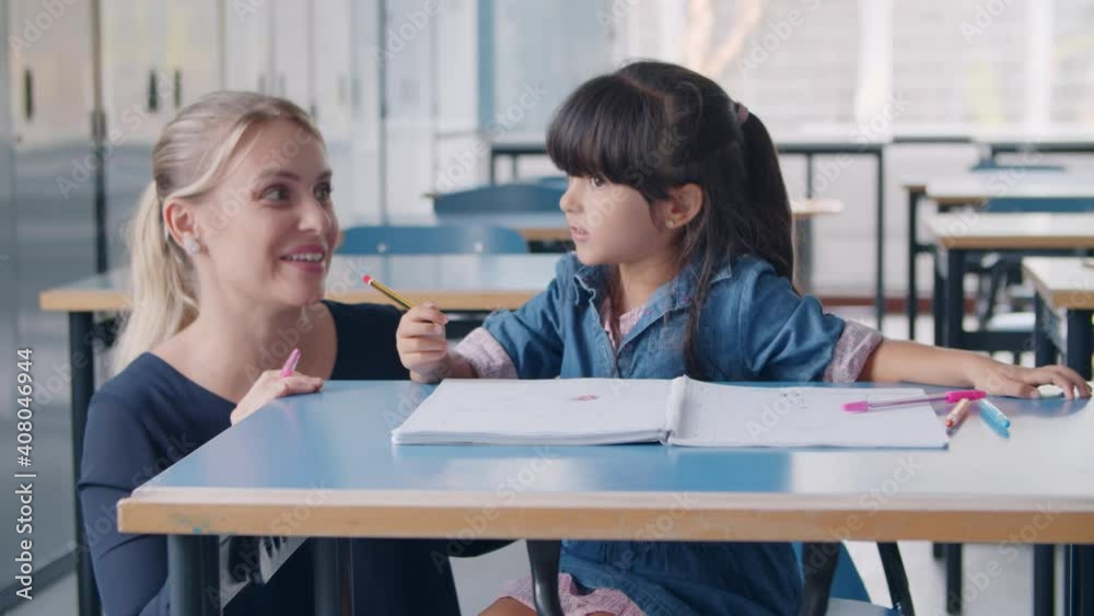 Happy female school teacher and Latin schoolgirl discussing task. Primary schoolkid holding pen, sitting at desk with copybook and speaking. Education or teaching concept Happy female school teacher and Latin schoolgirl discussing task. Primary schoolkid holding pen, sitting at desk with copybook and speaking. Education or teaching concept
