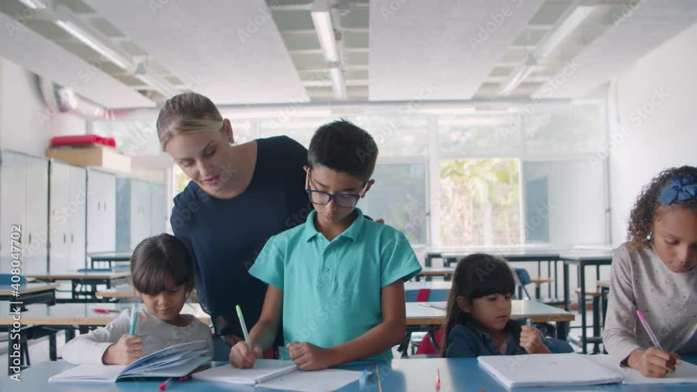 Female school teacher checking task of diverse group of pupils writing in copybooks at shared desk in classroom. Dolly shot. Education or teaching concept Female school teacher checking task of diverse group of pupils writing in copybooks at shared desk in classroom. Dolly shot. Education or teaching concept