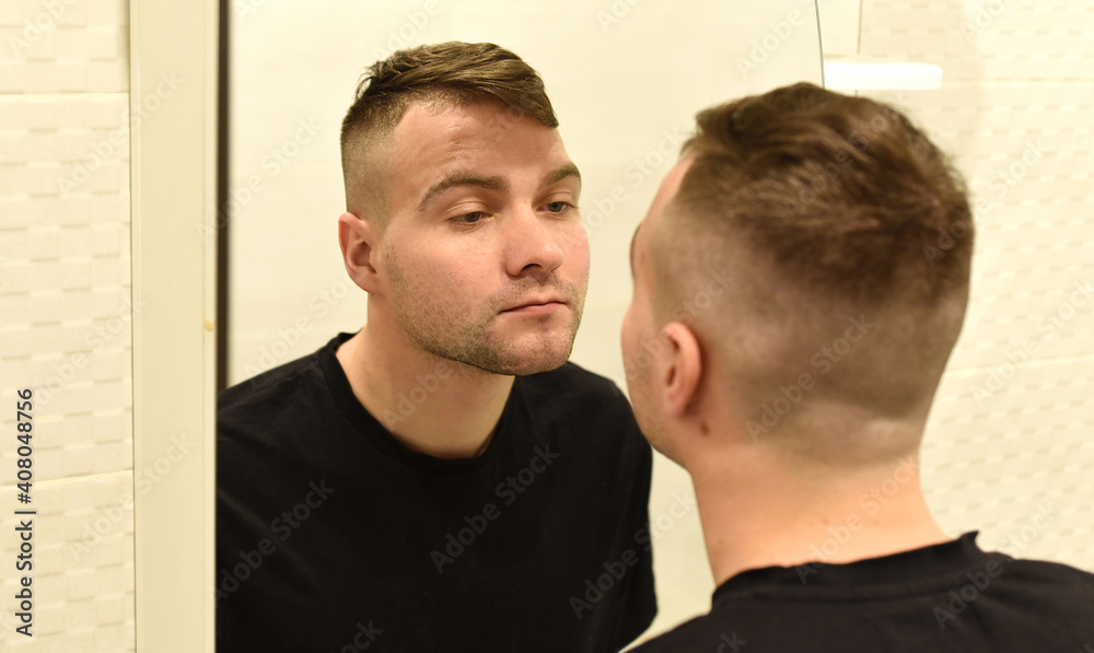 Young man looking in the mirror at his reflection in a bathroom Young man looking in the mirror at his reflection in a bathroom