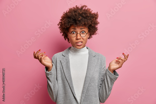 Photo of puzzled young African American woman entrepreneur with curly hair raises hands shrugs shoulders looks clueless or nervous says so what dressed in grey clothes poses against pink background