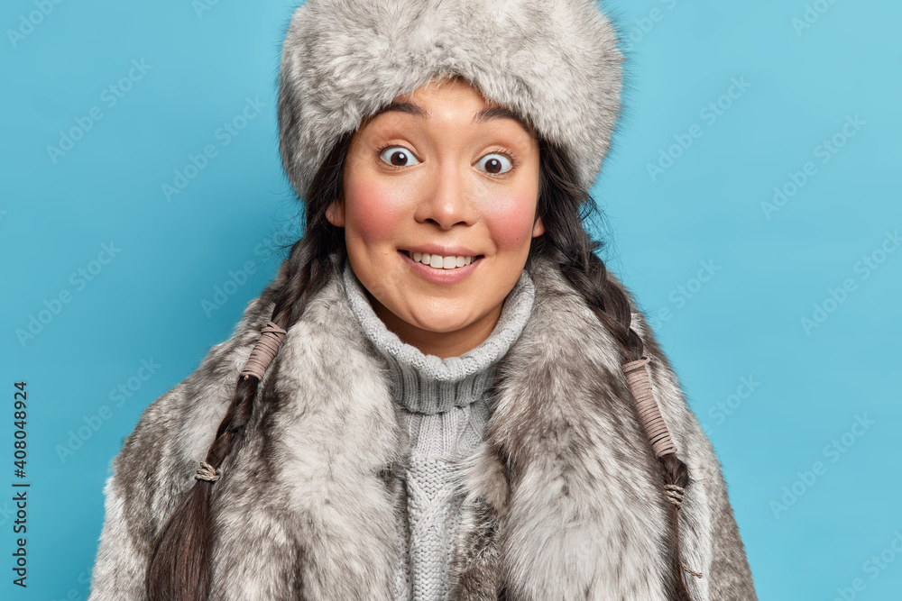 Close up shot of surprised Asian woman with two pigtails dressed in fur grey outerwear smiles pleasantly lives in arctic place isolated over blue background. Eskimo girl has walk during winter Close up shot of surprised Asian woman with two pigtails dressed in fur grey outerwear smiles pleasantly lives in arctic place isolated over blue background. Eskimo girl has walk during winter