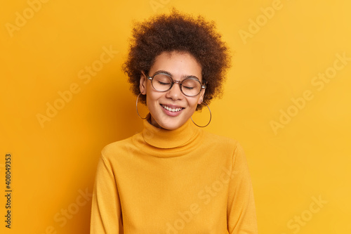 Horizontal shot of happy sincere millennial girl with curly hair natural beauty smiles gently with closed eyes gets compliment enjoys life wears casual poloneck isolated over yellow background © Wayhome Studio