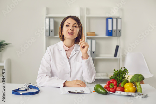 Young woman doctor nutritiologist sitting and speaking about healthy balanced diet for weight loss online looking at camera with various healthy ingredients on table. Healthy lifestyle concept © Studio Romantic