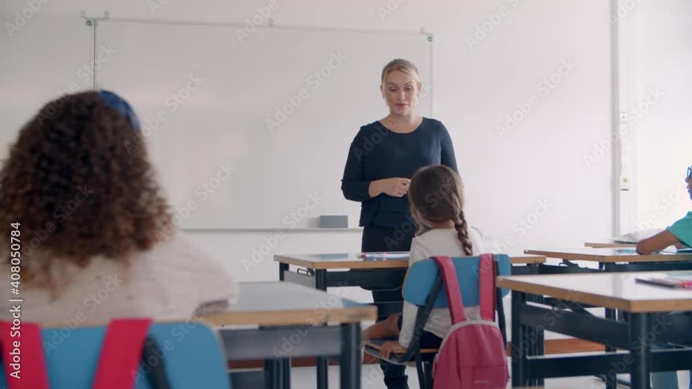 Friendly female school teacher speaking at whiteboard, explaining task to pupils in class, talking to kids. Dolly shot, back view. Teaching or education concept Friendly female school teacher speaking at whiteboard, explaining task to pupils in class, talking to kids. Dolly shot, back view. Teaching or education concept