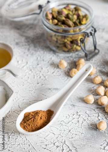 Closeup view of a ceramic white spoon full of ground cinnamon next to some hazelnuts over a rustic grey background.