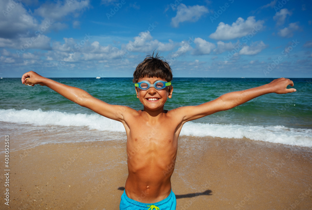 Happy smiling boy in swimming googles on the beach lift hands and smile laugh with sea on background Happy smiling boy in swimming googles on the beach lift hands and smile laugh with sea on background