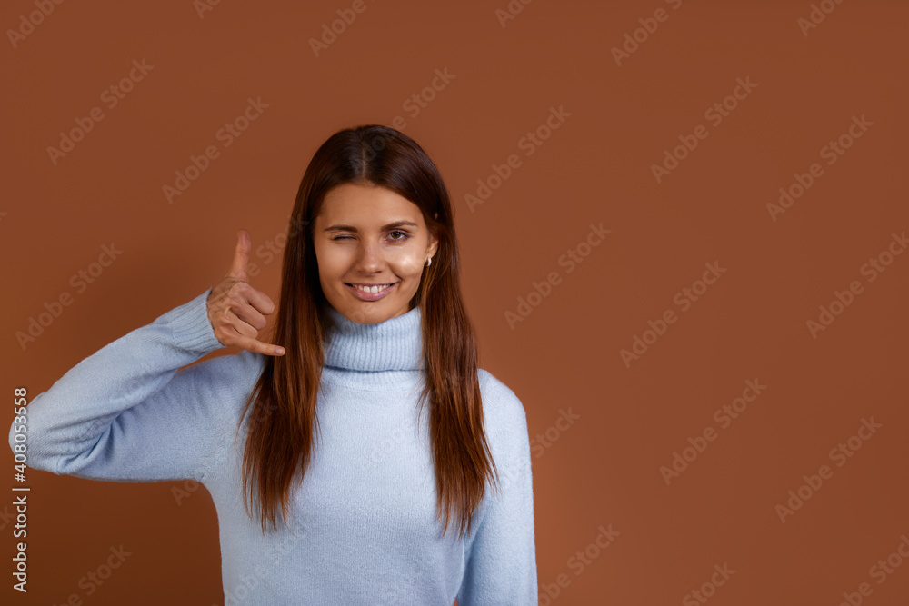 Call me back. Smiling caucasian woman wearing blue sweater makes telephone gesture, asks to call her, expresses positive emotions, blinks eyes, copy space on the right, isolated on brown background. Call me back. Smiling caucasian woman wearing blue sweater makes telephone gesture, asks to call her, expresses positive emotions, blinks eyes, copy space on the right, isolated on brown background.