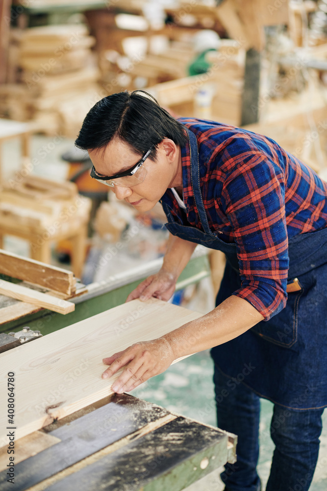Serious carpenter in apron and goggles working with industrial tool in wood factory and cutting wooden board with circular blade Serious carpenter in apron and goggles working with industrial tool in wood factory and cutting wooden board with circular blade