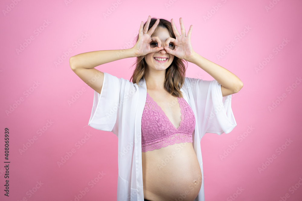 Young beautiful brunette woman pregnant expecting baby over isolated pink background doing ok gesture shocked with smiling face, eye looking through fingers Young beautiful brunette woman pregnant expecting baby over isolated pink background doing ok gesture shocked with smiling face, eye looking through fingers