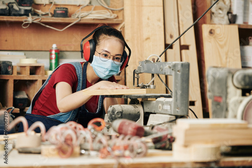 Tableau sur toile Concentrated young Asian carpenter in earmuffs and medical mask cutting wooden p