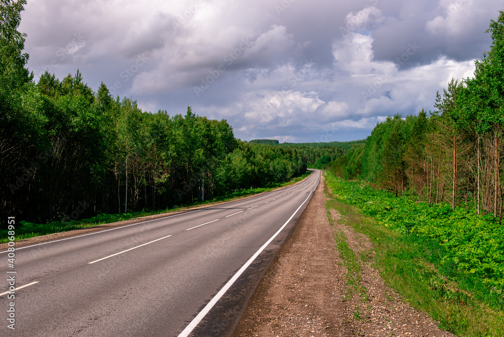 An asphalted intercity road with white markings extending into the distance through a green forest. An asphalted intercity road with white markings extending into the distance through a green forest.