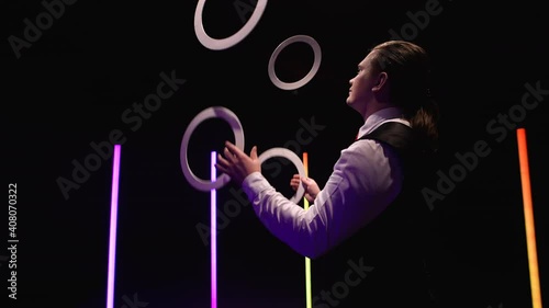 Camera rotates around professional male juggler juggling white rings. Stylish man hones his skill and reaction against black studio background amidst bright neon lights. Close up. Slow motion.