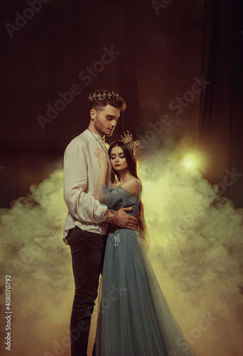Fantasy couple hugging in dark room full white smoke. Image of lovers king and queen. Medieval male prince in golden crown, vintage costume clothing. Girl Princess in long glamorous blue dress gown photo