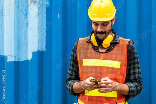 Caucasian worker in safety vest reflective with Safety helmet Typing a message on a cell phone in container pepot