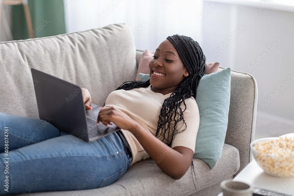 Joyful african lady laying on couch at home, using laptop Joyful african lady laying on couch at home, using laptop