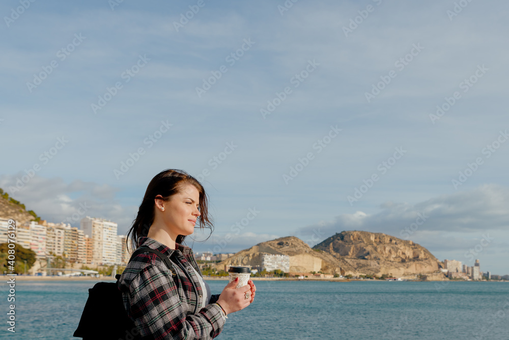 Traveler woman drinks coffee overlooking the sea and mountain landscape. A young tourist drinks a hot drink from a mug and enjoys the scenery of the city and the sea. Trekking concept