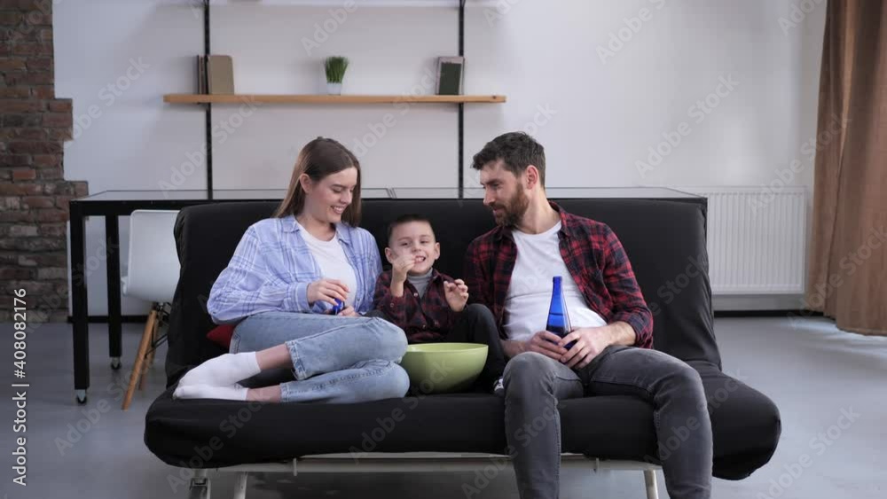 Family of three father mother little son watching TV at home. Young family getting positive emotions spending time together laughing hugging eating popcorn on the sofa. Togetherness. Stay home. Family of three father mother little son watching TV at home. Young family getting positive emotions spending time together laughing hugging eating popcorn on the sofa. Togetherness. Stay home.