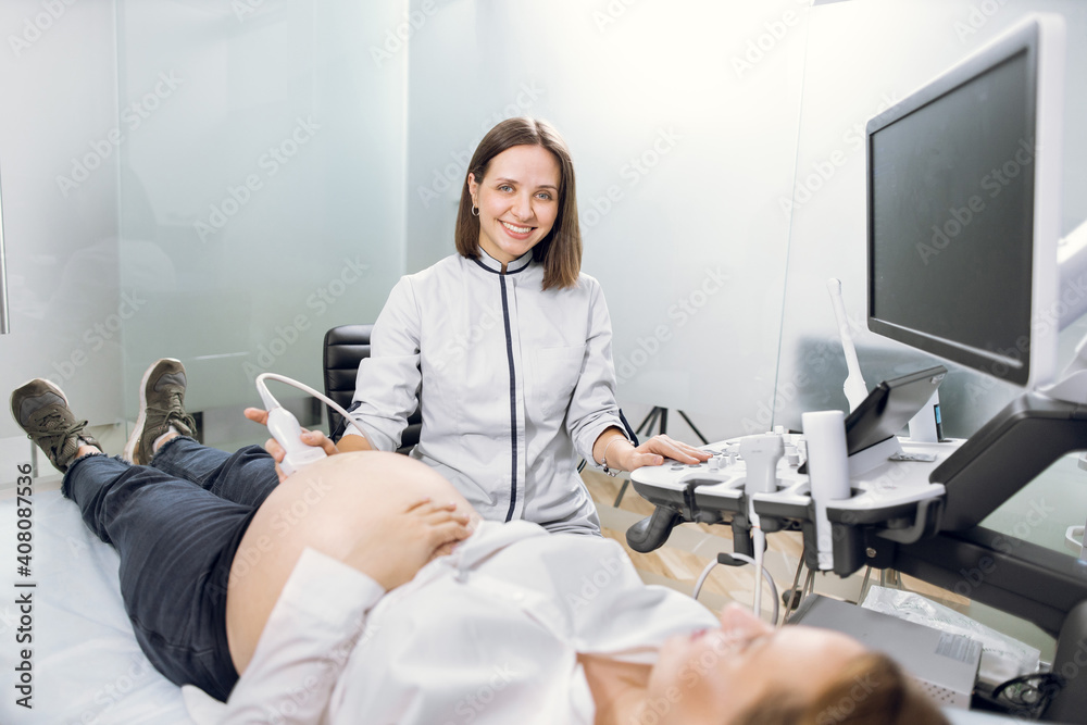 Obraz premium Portrait of young attractive female doctor, posing to camera with smile while conducting ultrasound scanning procedure for her patient, young pregnant woman. Ultrasound scan concept