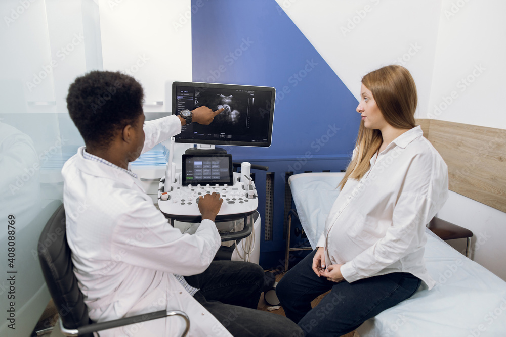 Shot of a beautiful young pregnant woman sitting on the couch in ultrasound examination room and having consultation with her male African doctor showing the results of ultrasound test on the screen