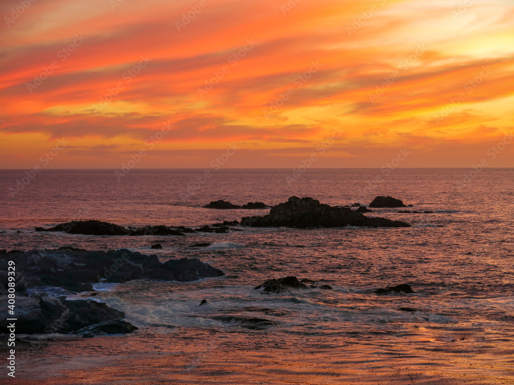 Sunset over Pacific Ocean near Carmel, California with reddish sky. Waves are hitting the rocks in the middle of the sea.