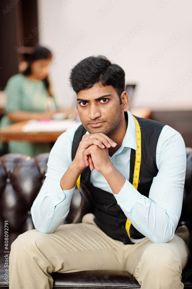 Portrait of young serious Indian tailor sitting on sofa in atelir and looking at camera Portrait of young serious Indian tailor sitting on sofa in atelir and looking at camera