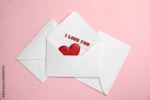 Sheet of paper with phrase I Love You and decorative heart in envelope on pink background, flat lay