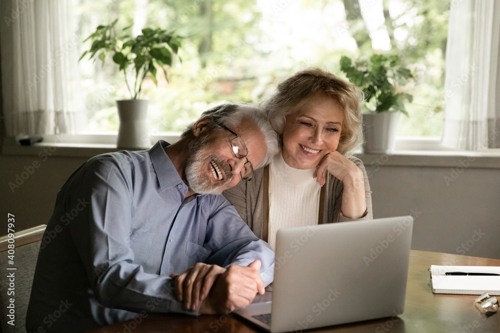 Happy mature man and woman hugging, looking at laptop screen, smiling elderly couple cuddling, watching movie or making video call to relatives, chatting online, enjoying leisure time with gadget Happy mature man and woman hugging, looking at laptop screen, smiling elderly couple cuddling, watching movie or making video call to relatives, chatting online, enjoying leisure time with gadget