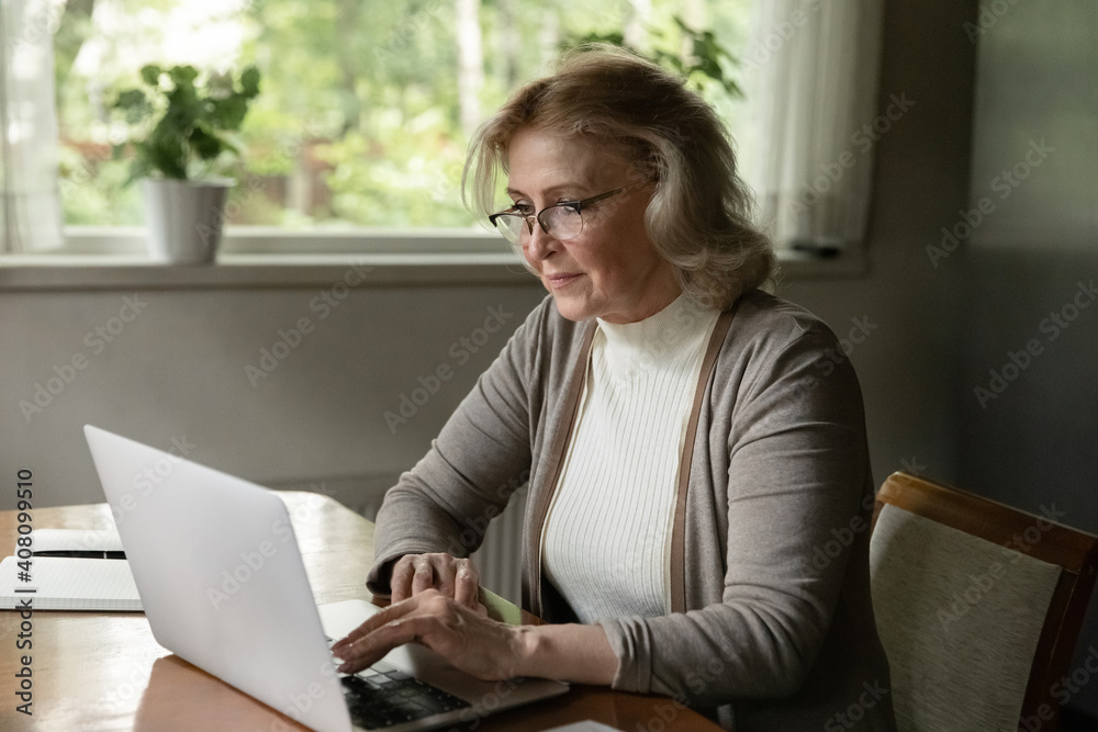 Confident mature woman wearing glasses using laptop, browsing apps, searching information in internet, shopping or chatting online, reading news, enjoying leisure time with gadget at home Confident mature woman wearing glasses using laptop, browsing apps, searching information in internet, shopping or chatting online, reading news, enjoying leisure time with gadget at home