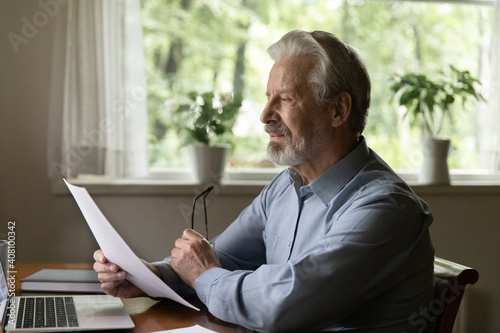 Focused elderly man reading letter, checking documents, working with correspondence, sitting at desk with laptop, taking off glasses, serious elderly male pondering strategy, analyzing expenses
