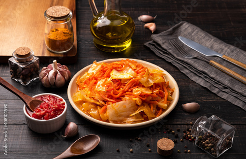 Spicy Korean appetizer carrot salad with cabbage on a plate surrounded by glass jars of spices on a dark wooden background. Horizontal orientation