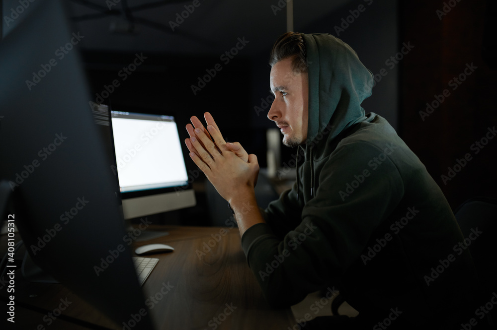 Young internet hacker in hood sitting at monitors