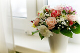 A bouquet of cut flowers in gentle tones of pink and white with green leaves stands on a windowsill against the background of a window in a vase