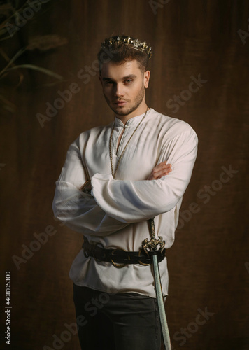 Portrait of handsome man in image of medieval king. Vintage clothing, historic white shirt retro clothing gold crown on head. Gothic room, studio. Fashion model posing. Adult strong sexy guy knight.