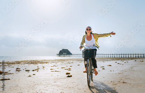 Young female dressed light summer clothes joyfully threw up her hand riding old vintage bicycle with front basket on the low tide ocean white sand coast on Kiwengwa beach on Zanzibar island, Tanzania. photo