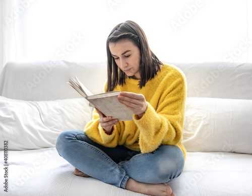 A cute girl in a yellow sweater is reading a book at home on the couch.