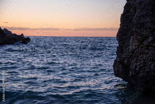 Sunset on sea coast with rocky cliffs. Seashore near a mountain with rocks and small waves. Closeup of stone rock in clear blue water.