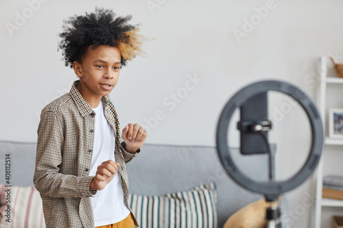 Waist up portrait of teenage African-American boy dancing to camera set on ringlight at home, young blogger concept, copy space photo