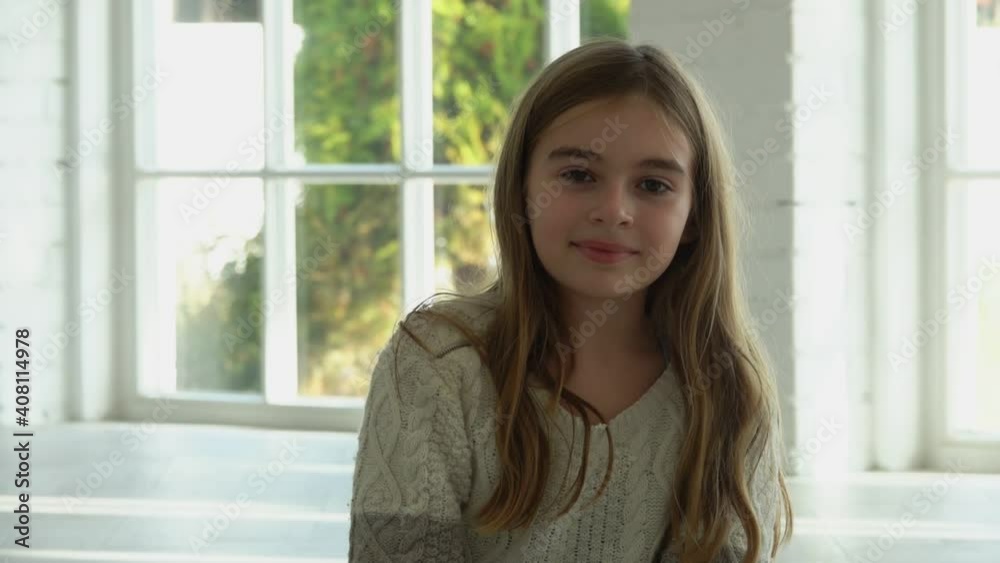 A beautiful little long-haired girl is smiling and sitting in a white sunlit hall with big windows A beautiful little long-haired girl is smiling and sitting in a white sunlit hall with big windows