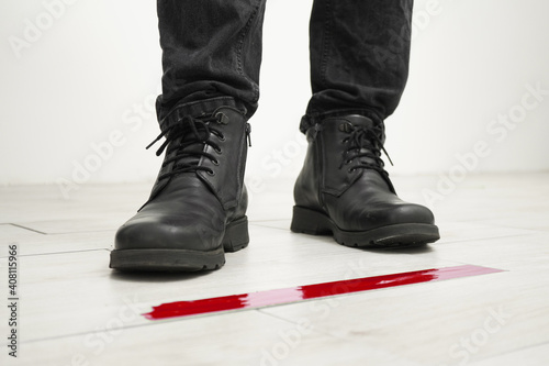 Man in black boots standing in line behind red tape floor marking for social distance indoors, close up. Social distancing for coronavirus prevention. Covid-19 pandemic concept.