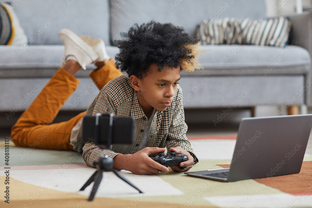 Full length portrait of teenage African-American boy playing videogames while lying on floor at home with camera filming for online streaming, copy space - obrazy, fototapety, plakaty 