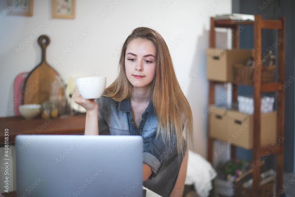 A young attractive girl with long hair has Breakfast, works and communicates on a laptop remotely in the kitchen at home.	