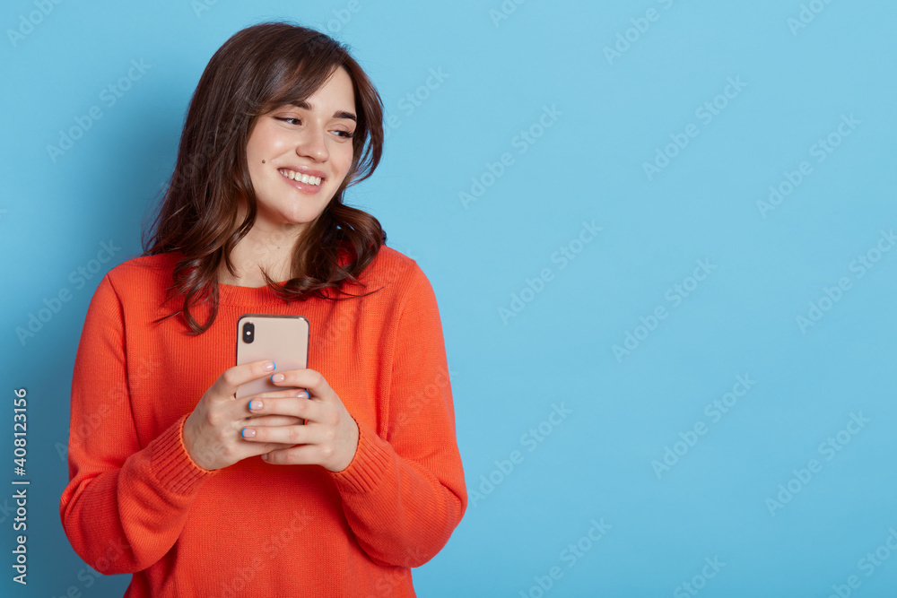 Carefree pleased female standing with happy look and holding mobile phone, looks aside and smiles broadly, browses through internet during free time, posing indoor, dresses casual attire. Carefree pleased female standing with happy look and holding mobile phone, looks aside and smiles broadly, browses through internet during free time, posing indoor, dresses casual attire.