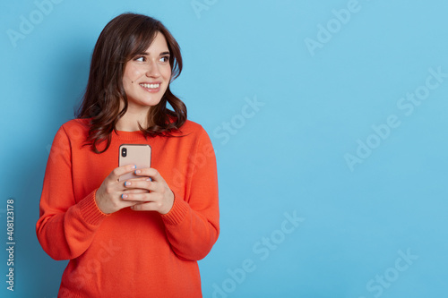 Pretty smiling woman holding smart phone, looks away with glad expression, uses mobile network service, posing isolated on blue background, copy space for advertisement.