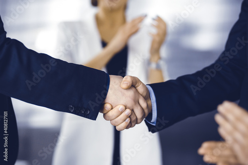 Businessmen are shaking hands while standing with colleagues applauding in office, close-up. Business success concept