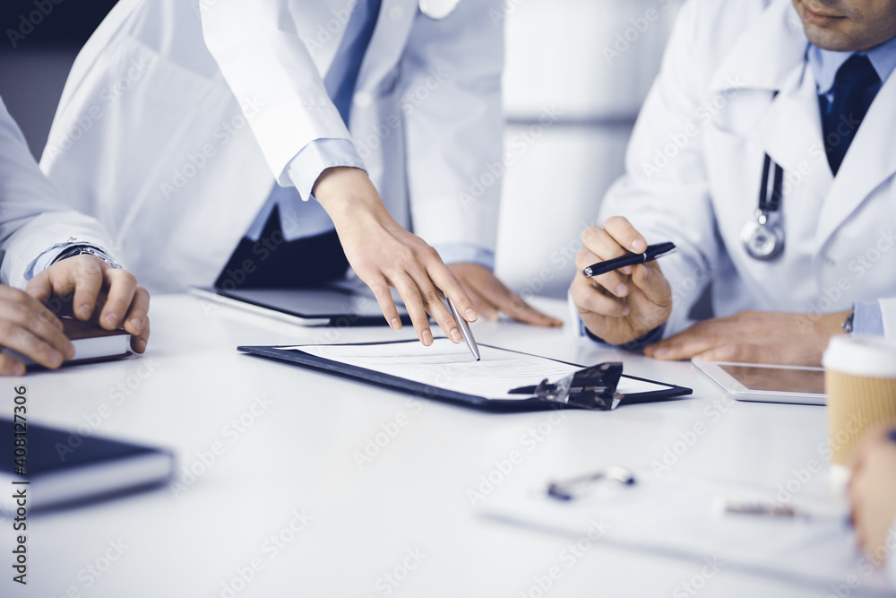 Group of unknown doctors are sitting at the desk and discussing medical treatment, using a clipboard, close-up. Team of physicians at work in a clinic. Medicine and healthcare concept Group of unknown doctors are sitting at the desk and discussing medical treatment, using a clipboard, close-up. Team of physicians at work in a clinic. Medicine and healthcare concept