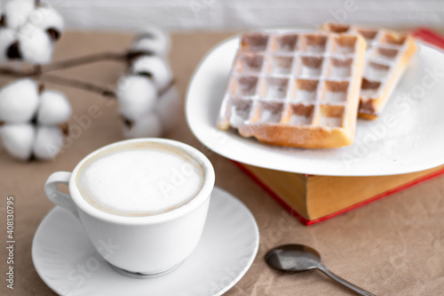 Viennese waffles on a white plate and a cup of cappuccino coffee laid out next to a branch of cotton
