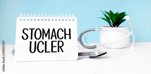The text STOMACH UCLER is written on notepad near a stethoscope on a blue background. Medical concept photo