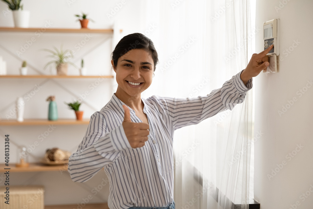 Portrait of smiling indian lady use domestic fire alarm system switch button on wall keypad show thumb up. Young female satisfied user of home security equipment look at camera trust recommend safety Portrait of smiling indian lady use domestic fire alarm system switch button on wall keypad show thumb up. Young female satisfied user of home security equipment look at camera trust recommend safety